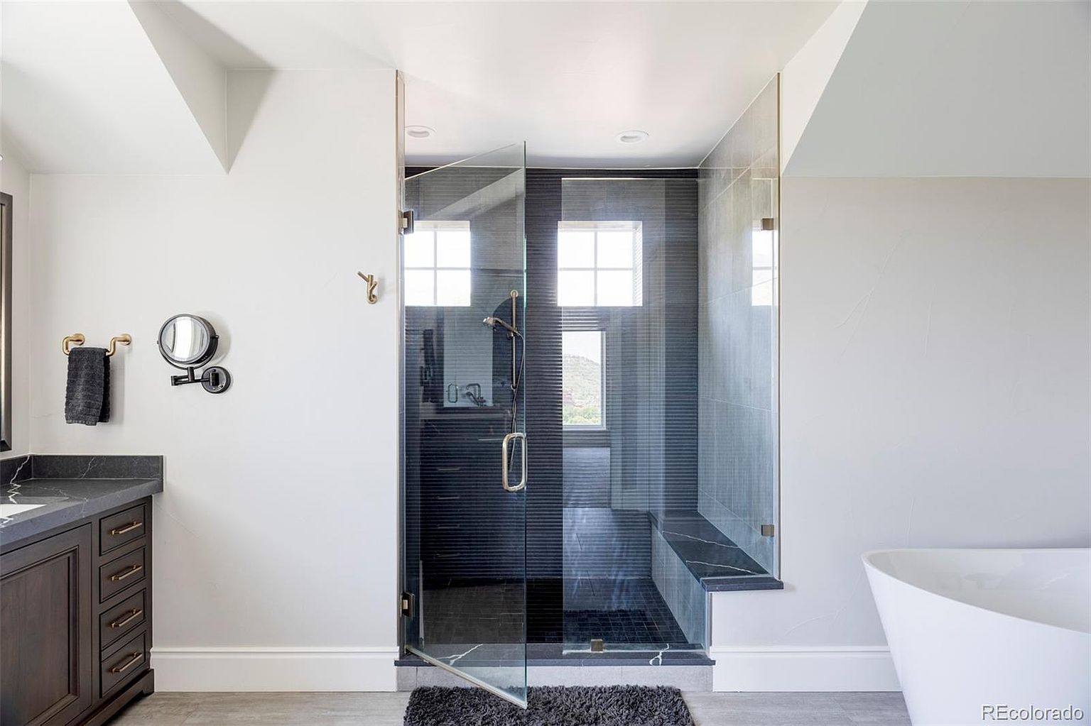 This is a well-lit primary bathroom featuring a modern glass-enclosed shower with dark tiling and a built-in bench. The bathroom includes a dark wood vanity with a black countertop, a round mirror, and gold fixtures, creating a sophisticated and luxurious feel. A freestanding white bathtub is partially visible on the right side of the image.