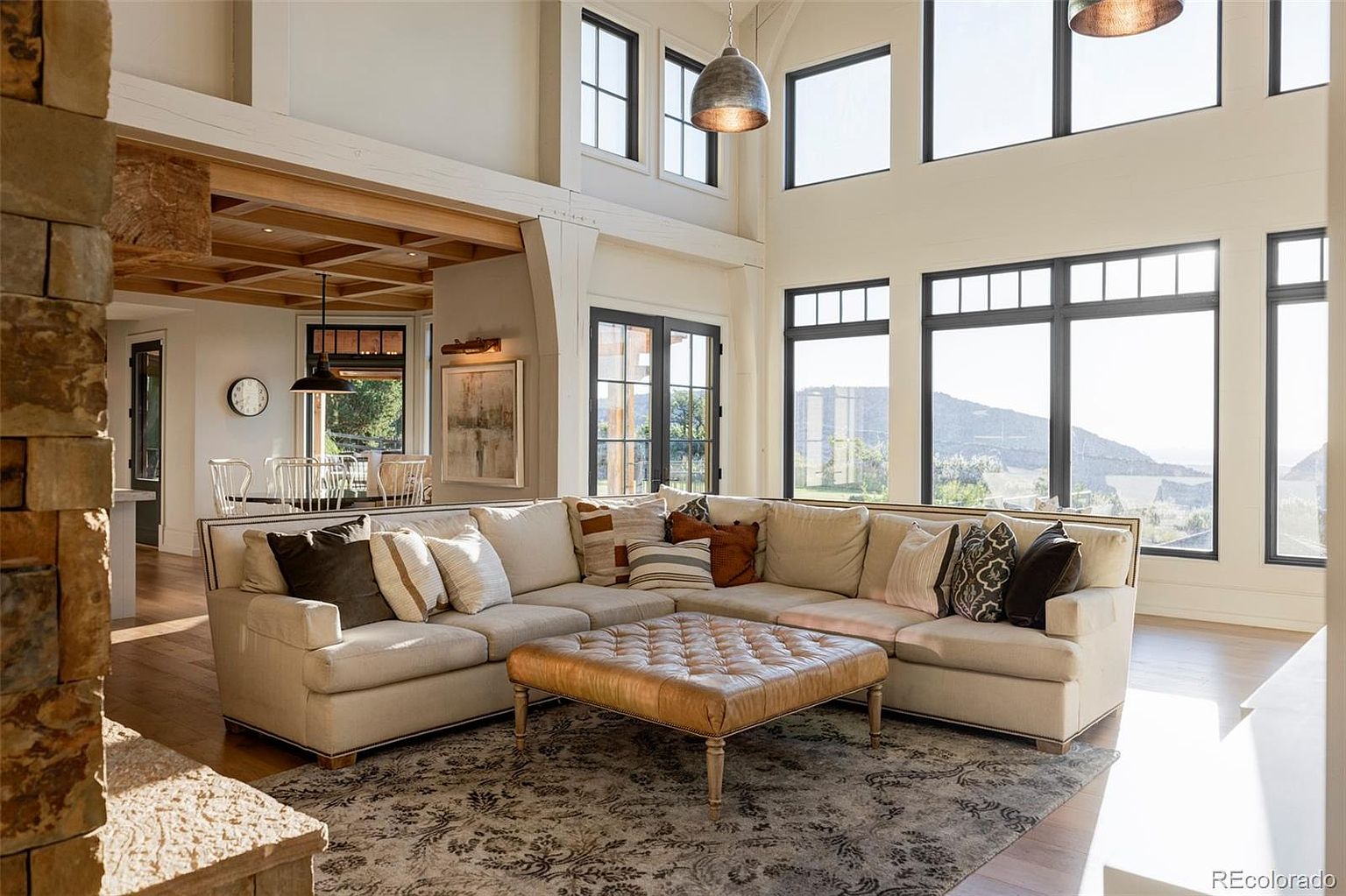 This is an interior shot of a luxurious living room featuring a large, comfortable sectional sofa with decorative pillows, a leather ottoman, and a patterned area rug. The room boasts high ceilings with large windows that provide ample natural light and scenic views. The design incorporates natural materials like wood and stone, creating a warm and inviting atmosphere.