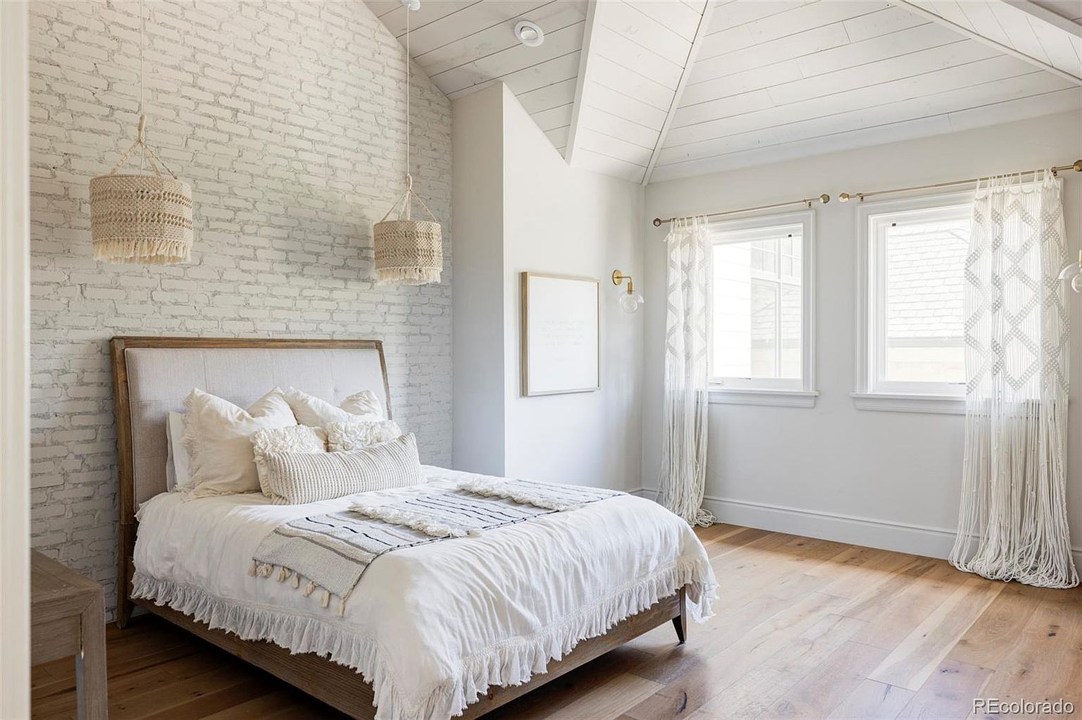 This is a bright and airy primary bedroom featuring a bed with a light-colored upholstered headboard and white bedding. The room has a vaulted ceiling with white wood paneling and two windows with sheer curtains. The walls are painted white, and the flooring is hardwood, creating a serene and inviting atmosphere.