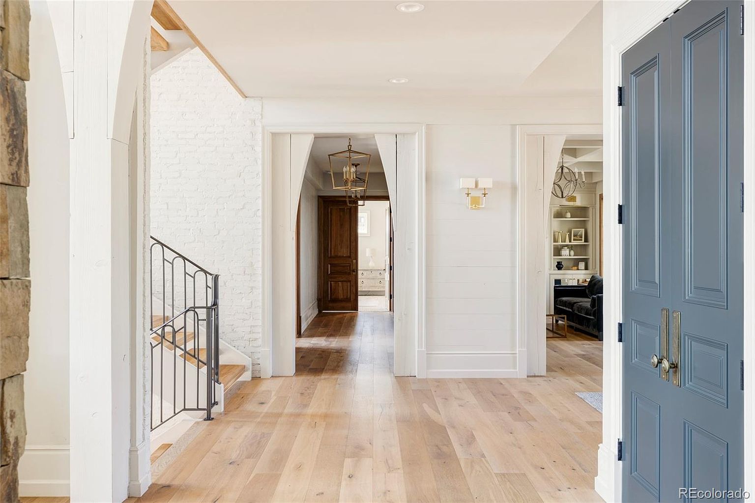 This interior shot showcases a bright and inviting hallway with light hardwood flooring and white walls. The entryway features a blue front door on the right, while a staircase with black iron railings is visible on the left. The hallway leads to other rooms, offering a glimpse of a living space with built-in shelves and a dark sofa, creating a sense of depth and spaciousness.