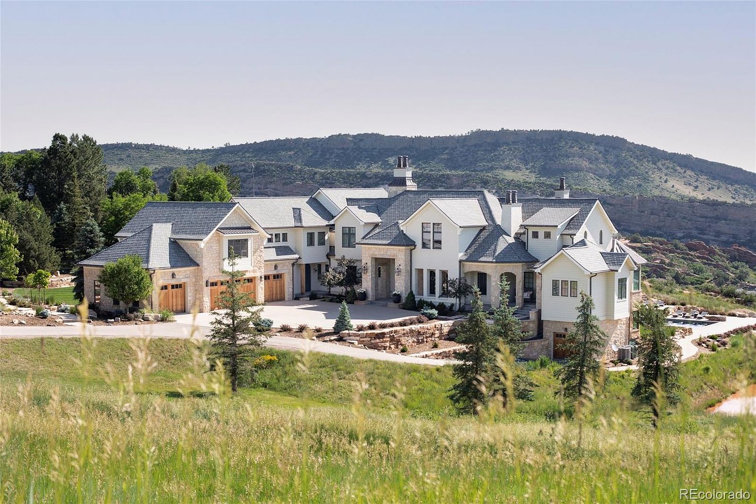 This is a stunning front view of a large, luxurious home. The house features a combination of stone and white siding, with a gray roof and multiple gables. A well-manicured lawn and landscaping add to the property's curb appeal, and a mountain range is visible in the background, creating a sense of privacy and grandeur.