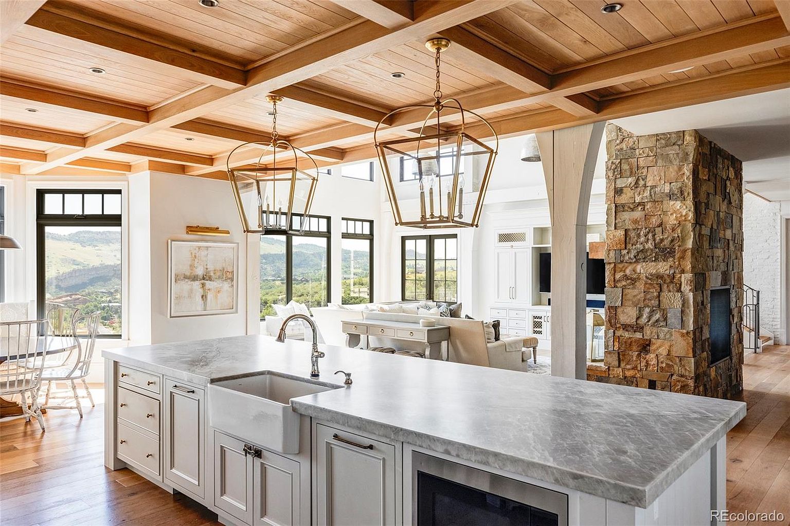 This is a bright and airy kitchen featuring a large island with a white farmhouse sink and marble countertops. The kitchen has custom white cabinetry and stainless steel appliances. The ceiling is wood paneled with exposed beams, and there are large windows offering views of the surrounding landscape. The kitchen opens into a living area with a stone fireplace.