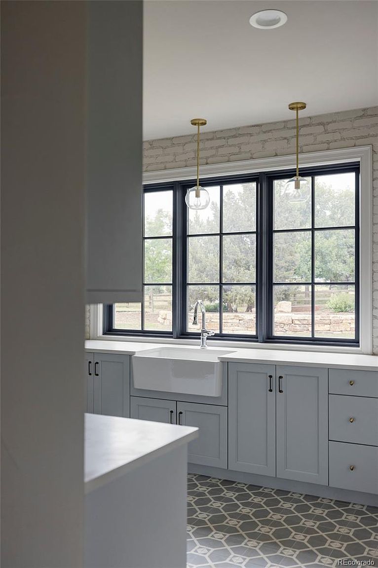 This is a well-lit kitchen featuring gray cabinetry, a white farmhouse sink, and black-framed windows offering a view of the outdoors. The countertops are light-colored, and the floor has a patterned tile design. Two pendant lights hang above the sink, adding a touch of elegance to the space.