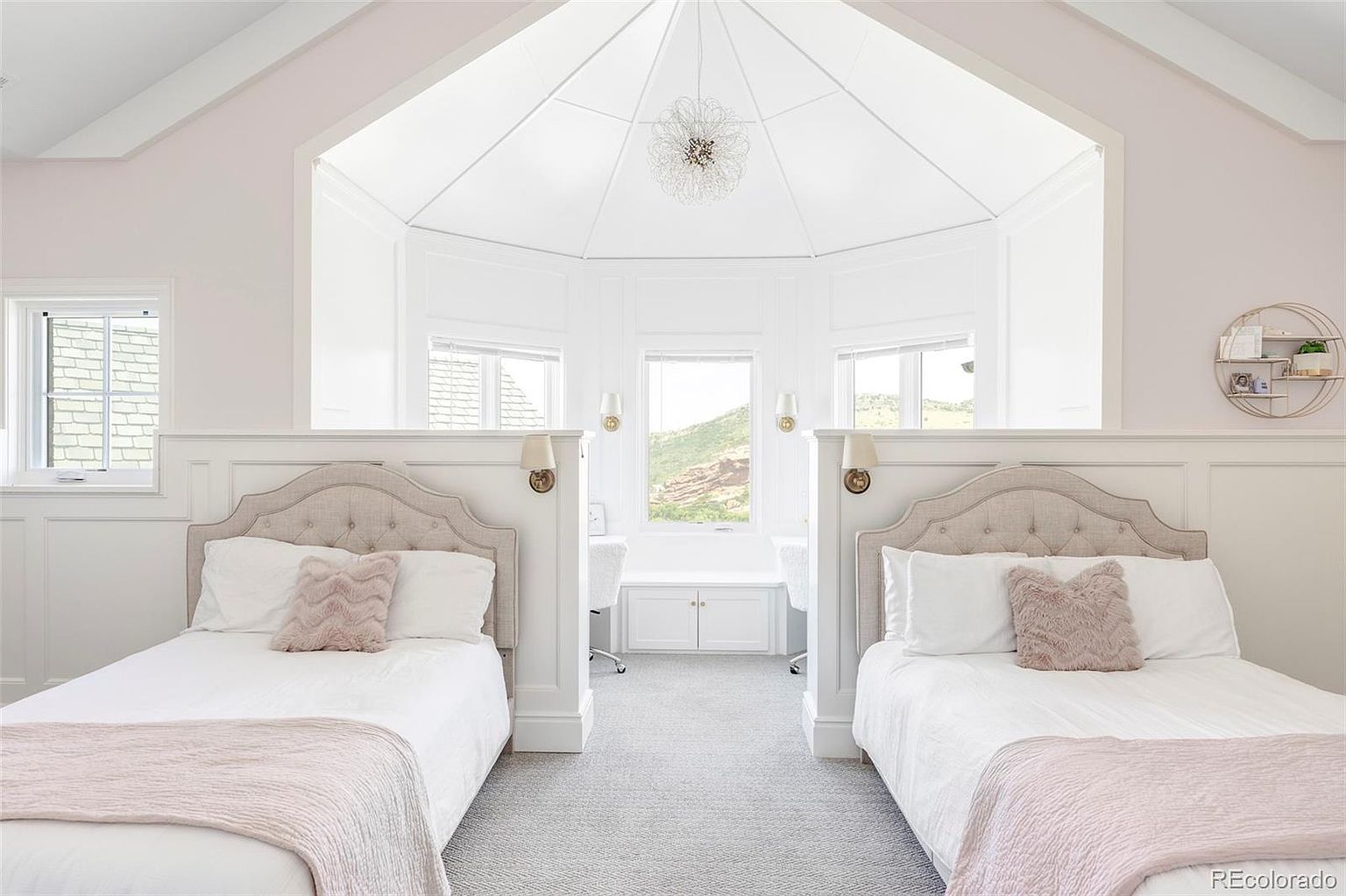 This is a bright and airy bedroom featuring two twin beds, each with a tufted headboard and soft pink accents. The room has a unique architectural design with a central bay window area offering views of the landscape. The overall aesthetic is clean, elegant, and inviting, perfect for a guest room or children's bedroom.