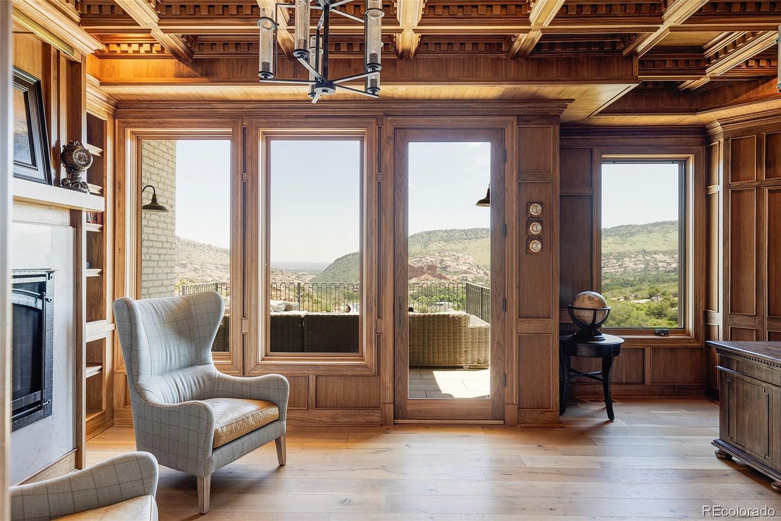 This is an interior shot of a luxurious living room featuring rich wood paneling and a coffered ceiling. Large windows offer a stunning view of the landscape, and comfortable armchairs invite relaxation. The room exudes a sense of warmth and sophistication, making it an ideal space for both entertaining and quiet enjoyment.