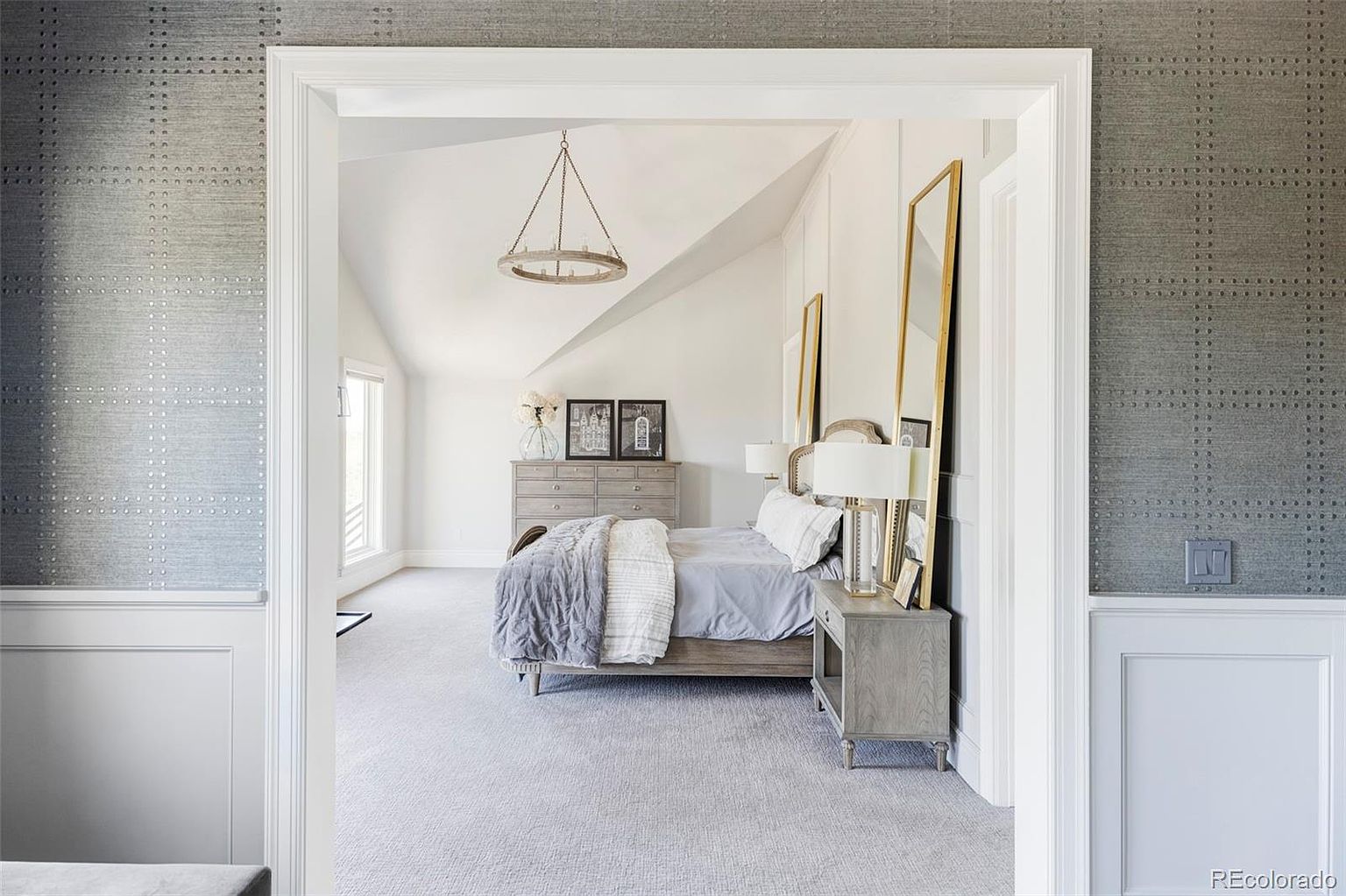 This is a view of a primary bedroom, seen through a doorway. The room features a neutral color palette with gray walls and carpet, a light wood bed frame, and a chandelier. There is a dresser against the far wall and tall mirrors flanking the bed, creating a bright and airy atmosphere.