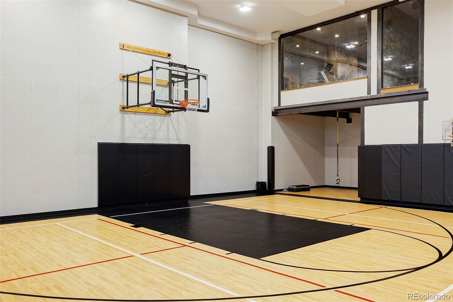 This is an indoor basketball court featuring a hardwood floor with painted lines and a black painted key area. A basketball hoop is mounted on the wall, and there is a glass-enclosed mezzanine area above, possibly for additional workout equipment. The space is well-lit and appears to be part of a larger residential property.