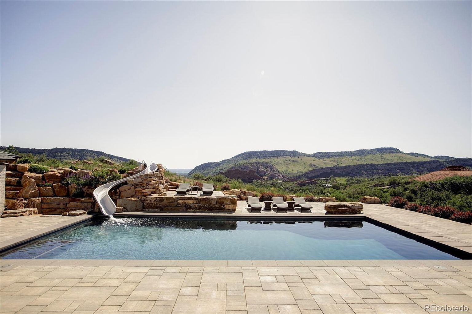 This image showcases a luxurious outdoor pool area with a slide and lounge chairs, set against a backdrop of rolling hills and a clear sky. The pool is surrounded by stone and paved areas, creating a sophisticated and relaxing environment. The presence of the slide adds a playful element to the upscale setting.