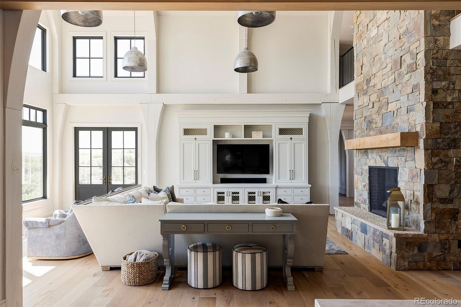 This is an interior shot of a living room featuring a stone fireplace, a large built-in entertainment center, and a neutral-toned sofa. The room has high ceilings with pendant lighting and large windows that provide natural light. The style is modern farmhouse with a focus on natural materials and a light color palette, creating a warm and inviting atmosphere.