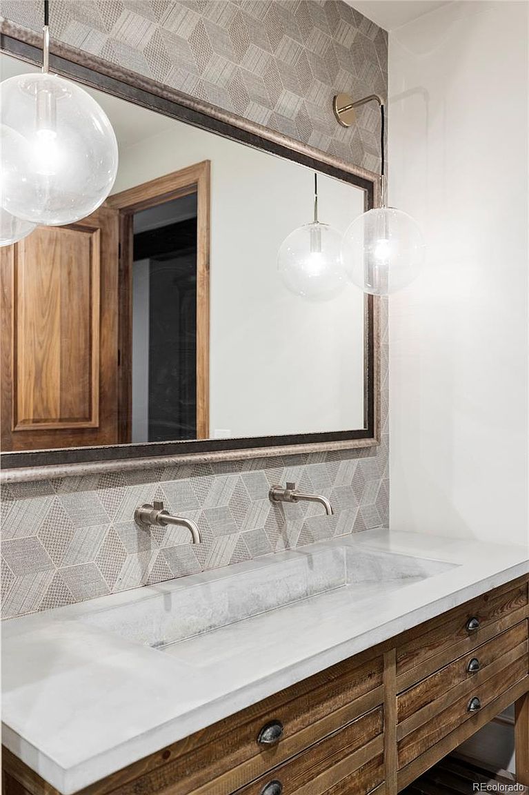 This is a stylish guest bathroom featuring a unique concrete sink with wall-mounted faucets and a rustic wooden vanity with drawers. A large mirror reflects the geometric patterned wallpaper and globe pendant lights, creating a modern yet warm aesthetic. The overall impression is sophisticated and well-designed.