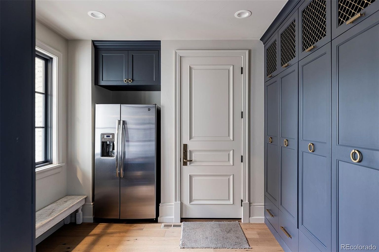 This interior shot showcases a well-organized laundry room featuring custom blue cabinetry with gold hardware, a stainless steel refrigerator, and a white paneled door. The room is illuminated by recessed lighting and natural light from a nearby window, creating a clean and functional space. The hardwood flooring adds warmth to the room.