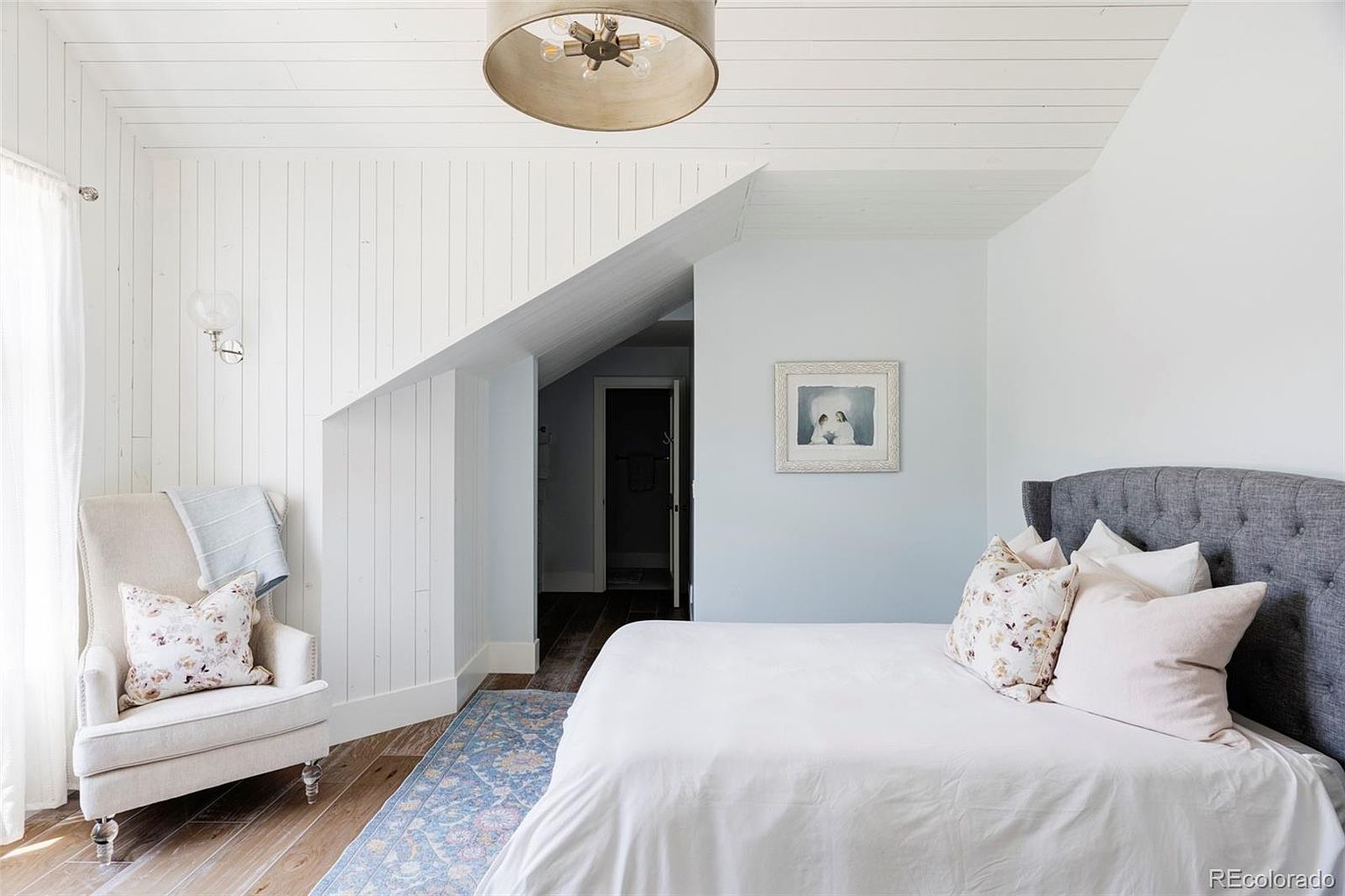 This is a cozy bedroom featuring a bed with a gray tufted headboard, white bedding, and decorative pillows. The room has white wood paneling on one wall and a light blue accent wall with a framed picture. A comfortable armchair sits near a window, and a blue patterned rug adds a touch of color to the hardwood floor. The room is well-lit with a unique ceiling light fixture.