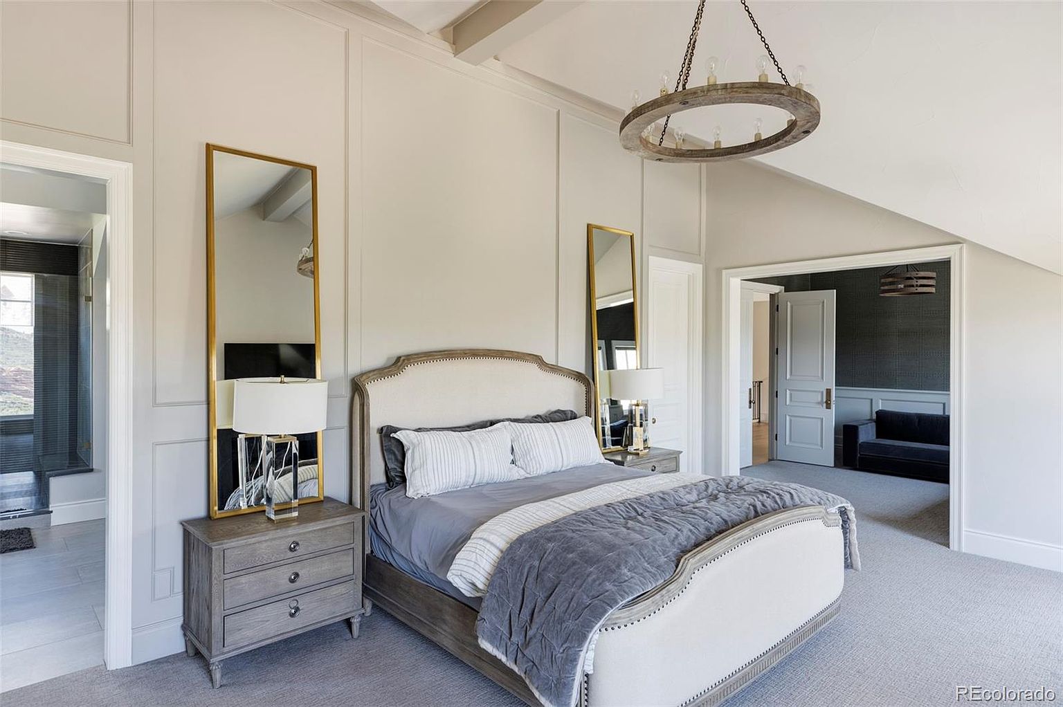 This is a primary bedroom featuring a neutral color palette with a large bed, two matching nightstands, and tall mirrors on either side. The room has a sophisticated and calming atmosphere, enhanced by the soft lighting from the lamps and the natural light filtering in. The space is well-appointed and designed for comfort and relaxation.