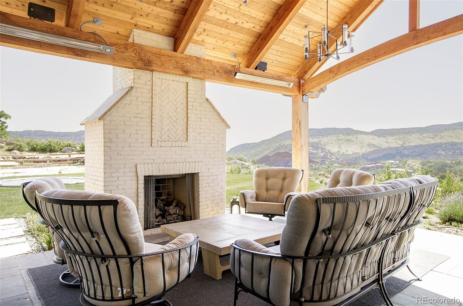 This image showcases a beautifully designed outdoor patio area, featuring comfortable seating arranged around a rustic coffee table and a charming brick fireplace. The space is covered by a wooden pergola, offering shade and architectural interest, while the background reveals a scenic mountain view, enhancing the property's appeal and creating a relaxing atmosphere.