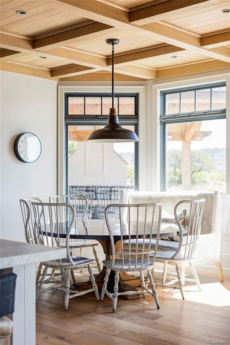 This is an interior shot of a dining room featuring a round table with a dark top and a light wood base, surrounded by light-colored chairs with spindle backs and a cushioned bench. Natural light floods the room through large windows, highlighting the wood-paneled ceiling and a modern pendant light fixture. A clock hangs on the wall, adding a touch of classic charm to the space.