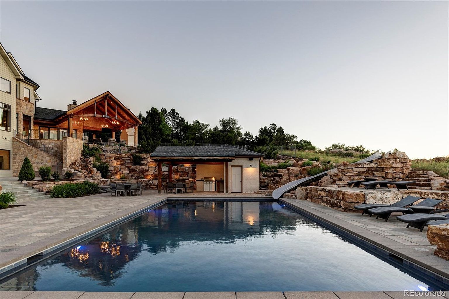 This image showcases a luxurious backyard with a stunning pool as the focal point. The pool is surrounded by stone patios, lounge chairs, and a built-in slide on a rock formation. A covered outdoor kitchen and dining area are adjacent to the pool, creating an ideal space for entertaining. The overall impression is one of high-end outdoor living and relaxation.