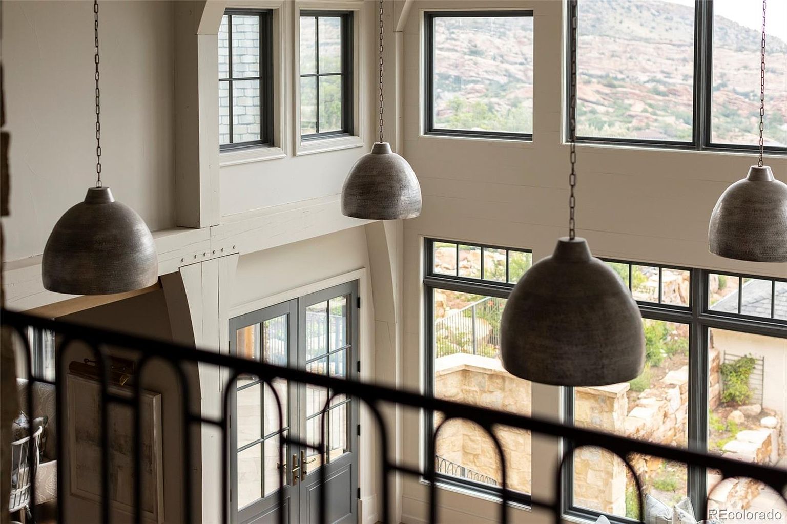 This interior shot captures a grand hallway or entryway from an elevated perspective, showcasing a double-door entrance below and large windows that flood the space with natural light. The design features elegant pendant lighting, a decorative railing, and neutral color tones, creating a bright and inviting atmosphere.