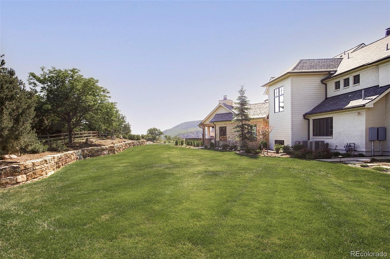 This image showcases a spacious and well-maintained backyard featuring a lush green lawn. A stone retaining wall adds character and definition to the landscape, while the house, with its combination of stone and siding, provides an elegant backdrop. The overall impression is one of tranquility and ample outdoor living space.
