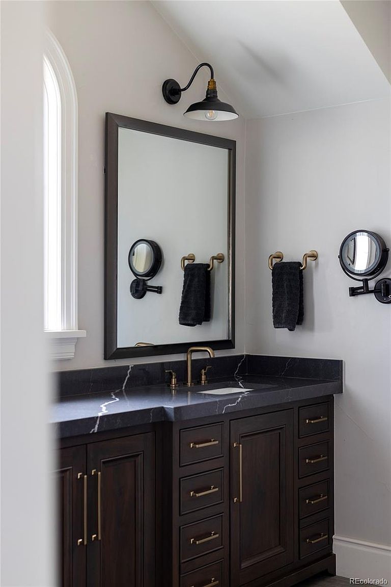 This is a well-appointed bathroom featuring a dark wood vanity with gold hardware and a black marble countertop. A large framed mirror hangs above the vanity, flanked by sconce lighting and decorative mirrors. The overall aesthetic is modern and sophisticated, with a focus on high-end finishes.