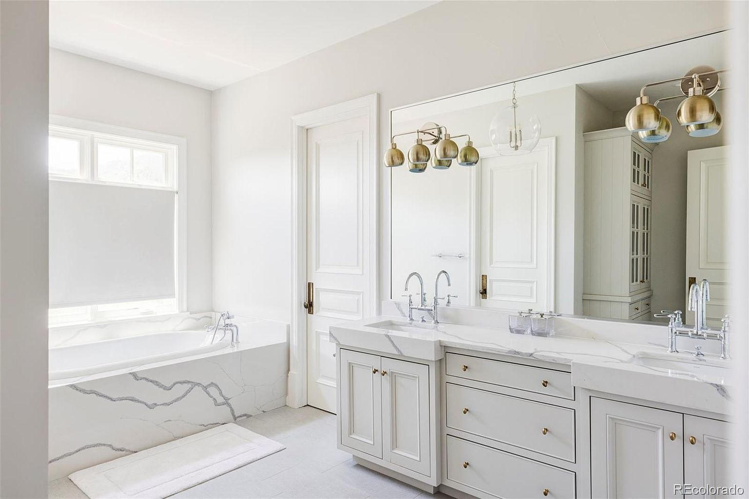 This is a bright and luxurious primary bathroom featuring a double vanity with white marble countertops and cabinetry. A large mirror reflects the space, enhancing the sense of openness, while a freestanding bathtub sits beneath a window, creating a spa-like atmosphere. The overall design is elegant and modern, with a focus on clean lines and high-end finishes.