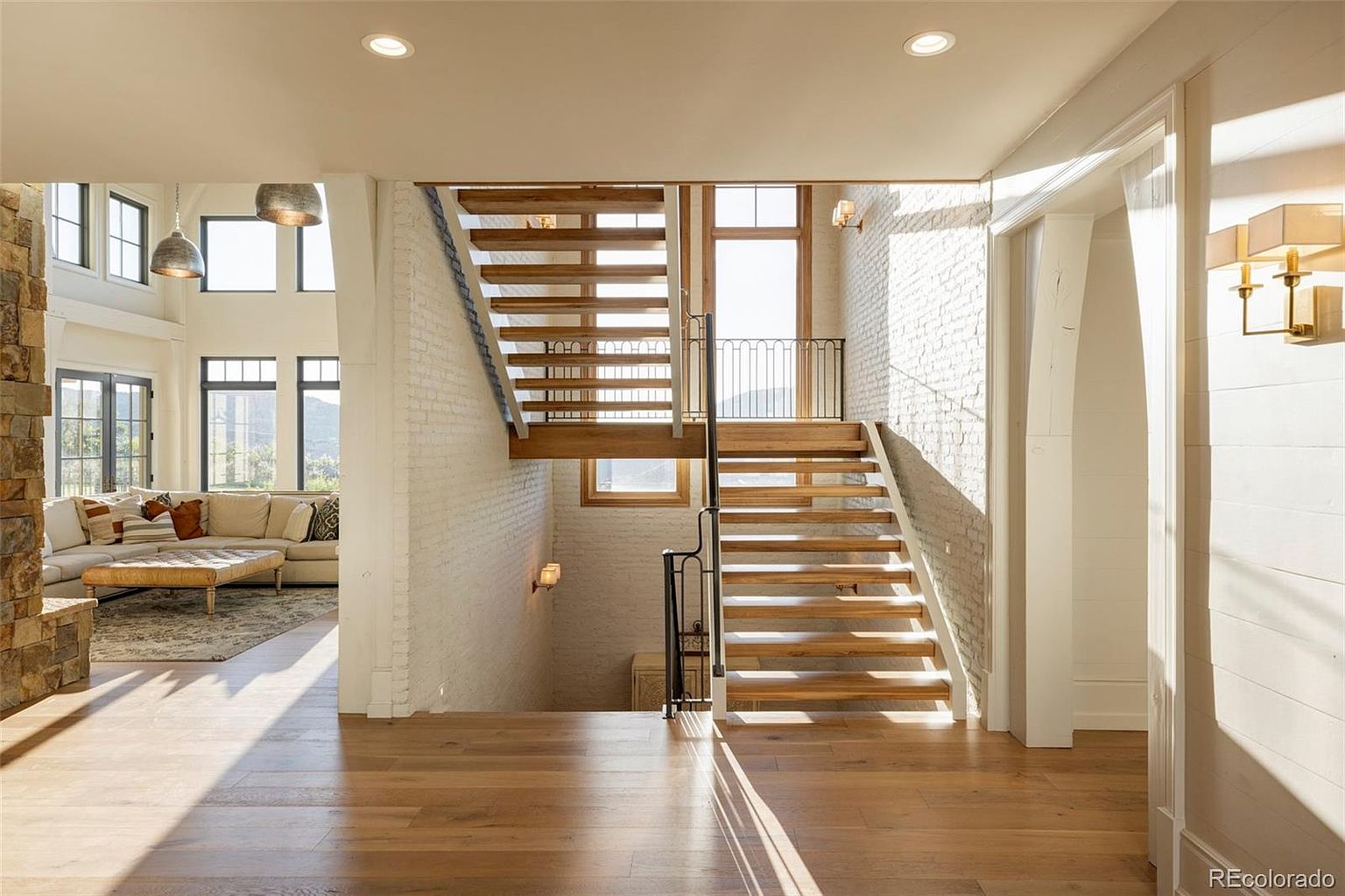 This interior shot showcases a modern hallway with striking architectural stairs. The stairs feature open wooden treads and a minimalist metal railing, set against a backdrop of white brick walls and natural light streaming through a window. The hardwood flooring adds warmth to the space, creating an inviting and stylish transition area.