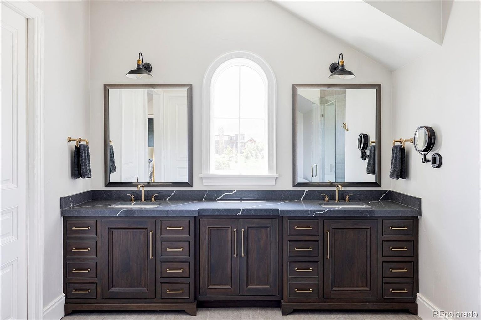 This is a well-lit primary bathroom featuring a double vanity with dark wood cabinetry and a dark countertop with light veining. Each sink has its own mirror and sconce lighting, and there is a large arched window between the mirrors. The overall style is modern and luxurious.