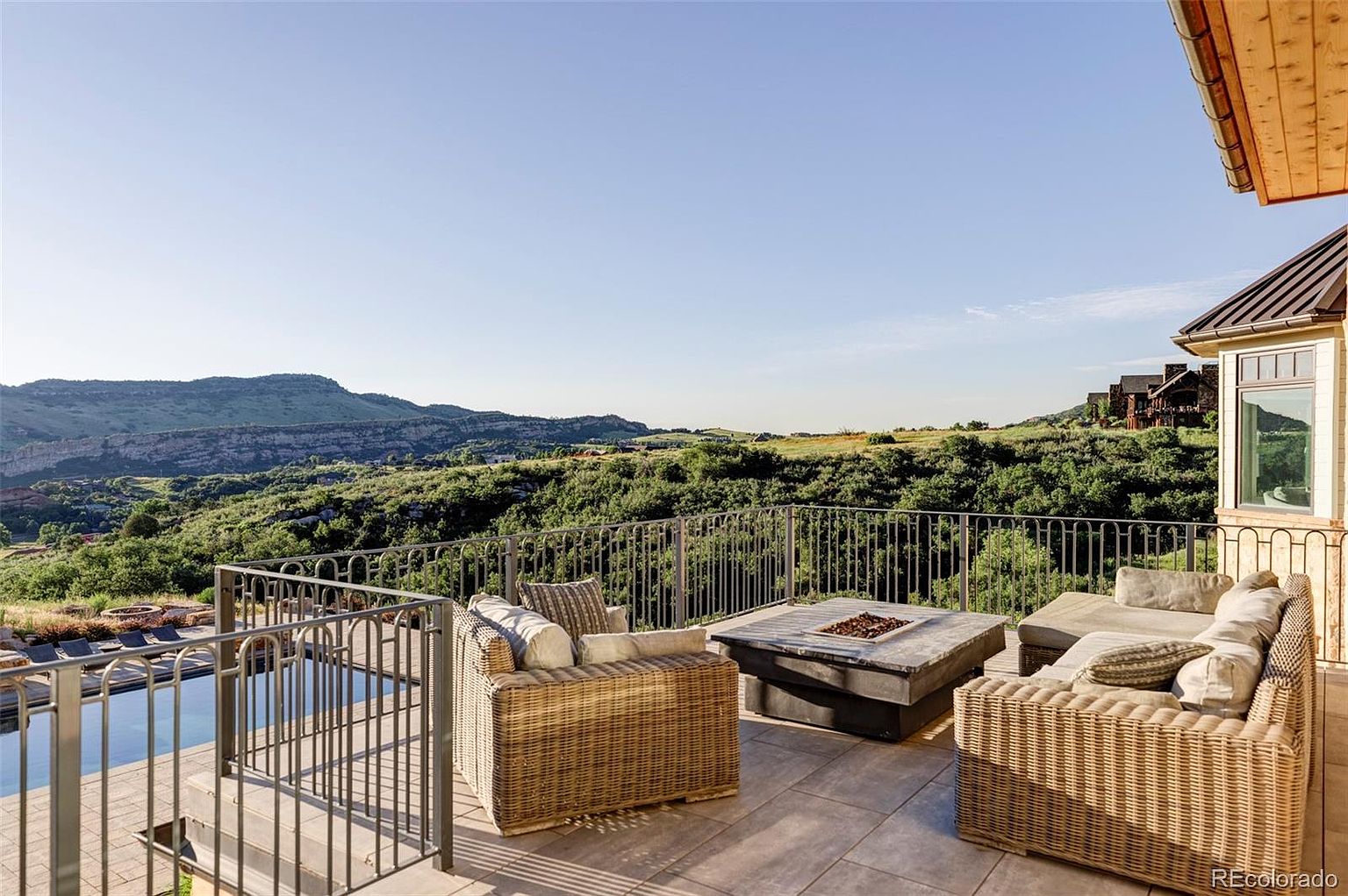 This image showcases a luxurious outdoor patio area with comfortable wicker seating arranged around a modern fire pit. The patio is elevated, offering stunning views of the surrounding landscape, including rolling hills and lush greenery. A sleek railing adds a touch of elegance and safety to the space, while a glimpse of a pool suggests additional outdoor amenities.