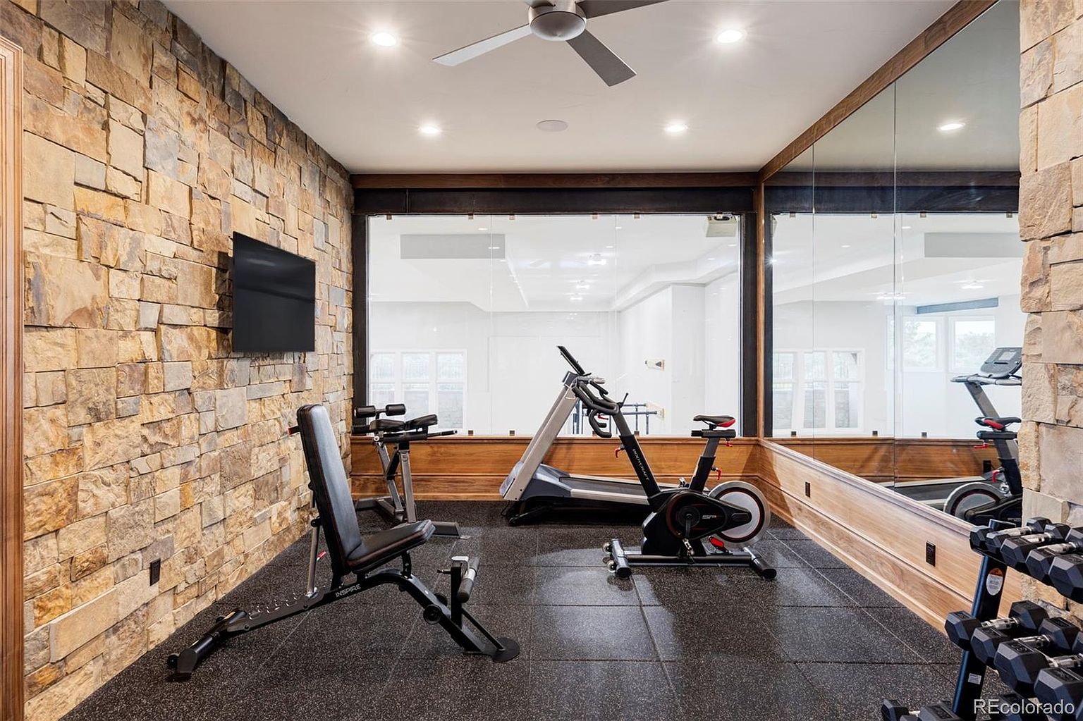 This is an interior shot of a home gym featuring stone accent walls and a mirrored wall. Exercise equipment such as a treadmill, stationary bike, and weight bench are visible. The room has a modern and functional design, perfect for a workout space.