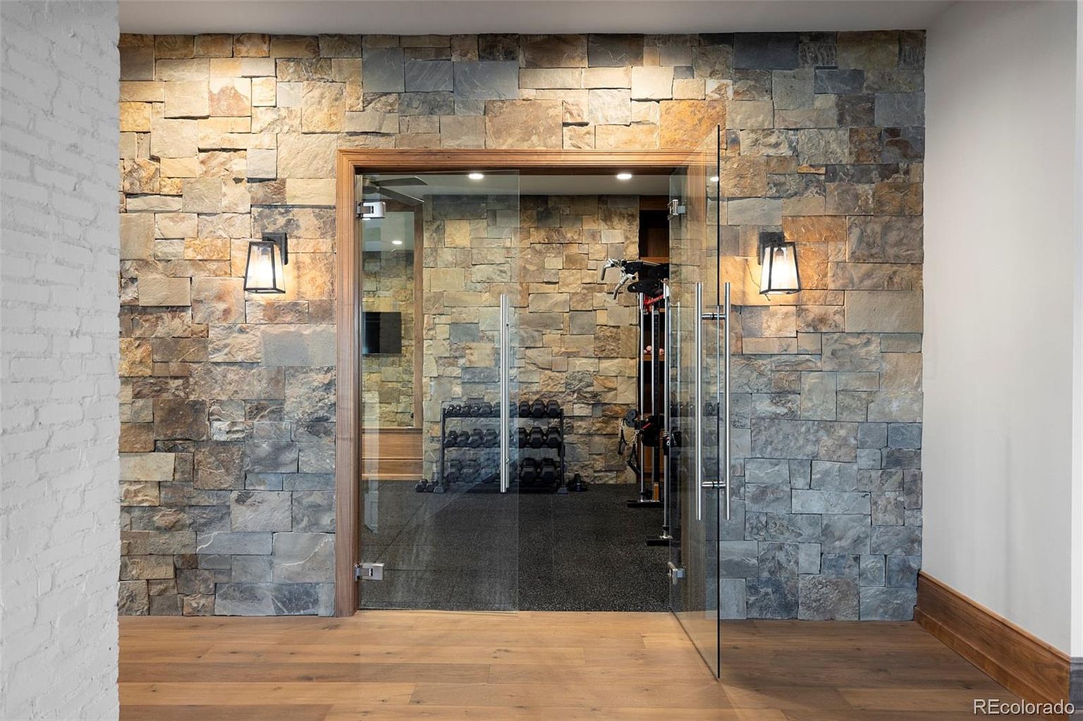 This image showcases a home gym with a stone accent wall and glass doors. The gym features weightlifting equipment and dumbbells, suggesting a dedicated space for fitness. The hardwood flooring and sconce lighting add a touch of elegance to the functional area.