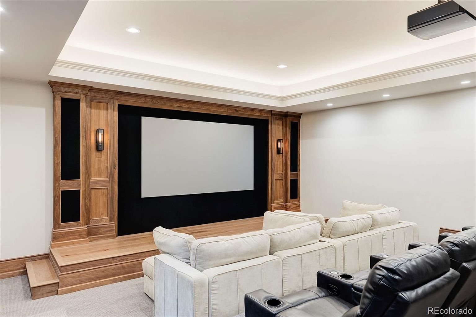This is an interior shot of a home theater room. The room features a large screen framed by wood paneling with sconce lighting, tiered seating with plush sofas and leather recliners, and a projector mounted on the ceiling. The overall impression is one of luxury and comfort, perfect for a cinematic experience.