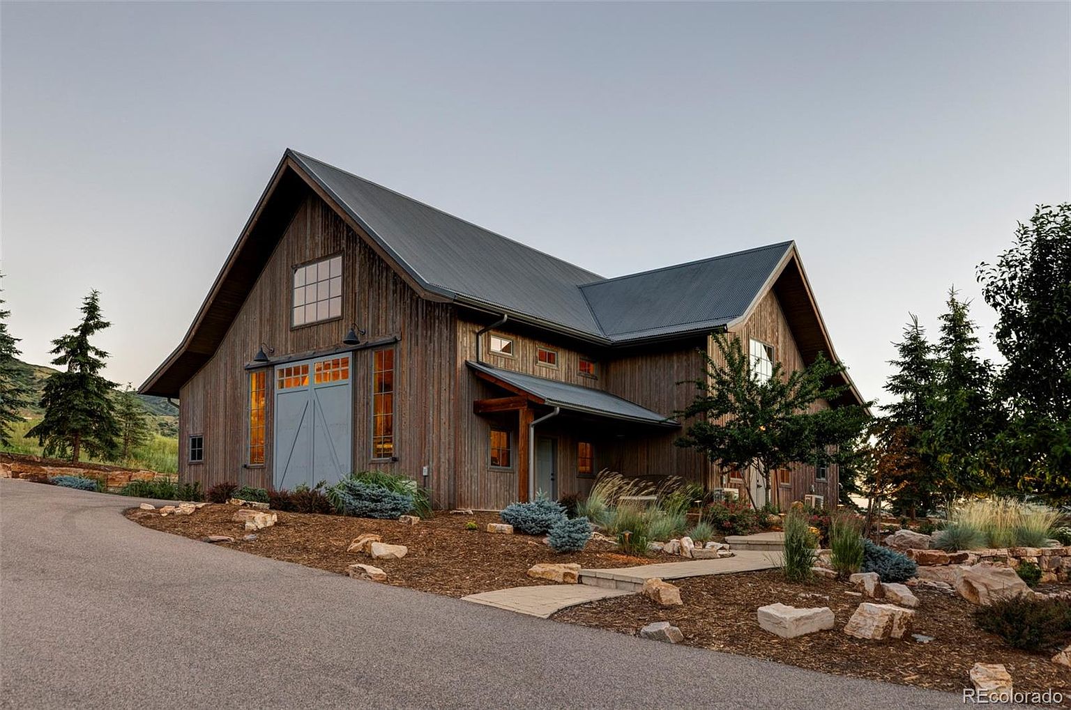 This is a front exterior view of a barn-style home with a metal roof and wooden siding. The property features a well-maintained landscape with rocks, mulch, and various plants. A paved driveway leads up to the house, enhancing its curb appeal and showcasing its unique architectural design.