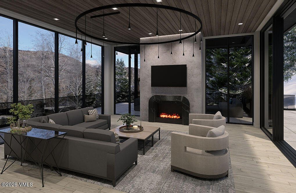 This is a modern living room featuring a gray sectional sofa, two armchairs, and a fireplace with a television mounted above. Large windows offer a view of the outdoors, and a circular lighting fixture hangs from the ceiling. The room is decorated in neutral tones with a contemporary style, creating a cozy and inviting atmosphere.