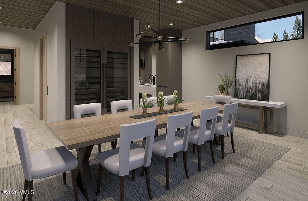 This is a dining room featuring a long wooden table with white chairs, set on a gray rug. A modern chandelier hangs above the table, and a console table with artwork is placed against the wall. The room has a contemporary design with a neutral color palette and a view through a horizontal window.