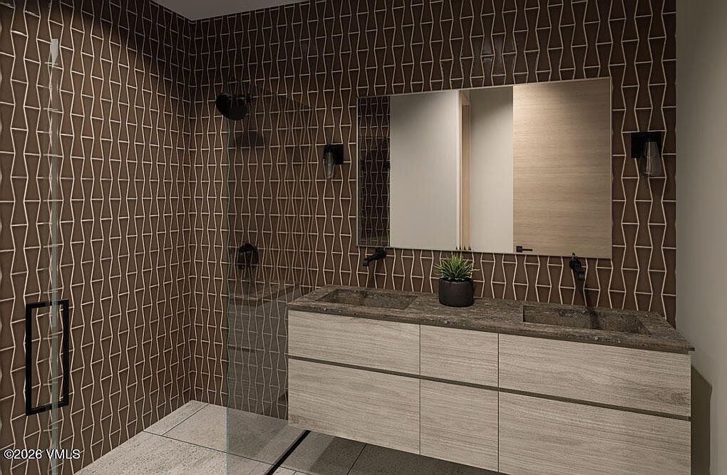 This is a modern primary bathroom featuring a double vanity with a light wood finish and dark stone countertops. The walls are covered in a unique brown tile pattern, complemented by black fixtures. A glass-enclosed shower is visible to the left, and a large mirror hangs above the vanity, enhancing the sense of space.