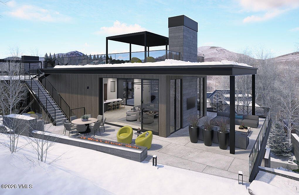 This image showcases a luxurious patio/deck area of a modern home, featuring outdoor seating, a fire pit, and sleek planters. The space is designed for relaxation and entertainment, with seamless indoor-outdoor flow through large glass doors. The architectural style is contemporary, emphasizing clean lines and high-end finishes, with a mountain view in the background.