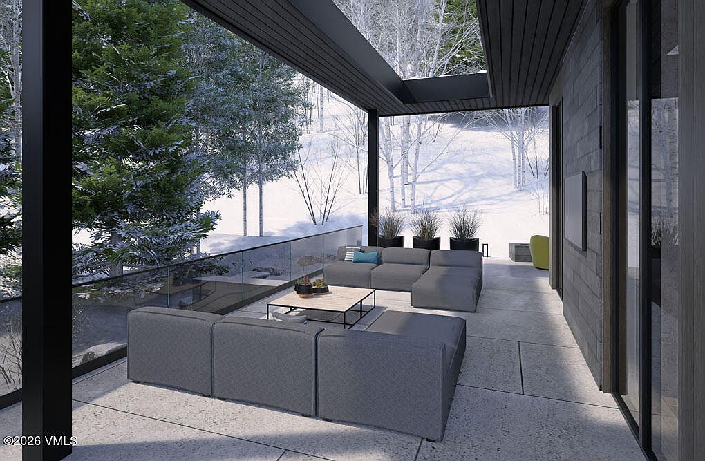 This image showcases a modern patio or balcony area with sleek, gray modular seating arranged around a low coffee table. The space is enclosed by a glass railing, offering unobstructed views of a snowy, wooded landscape. The architectural design features clean lines and a minimalist aesthetic, creating a serene and sophisticated outdoor living space.