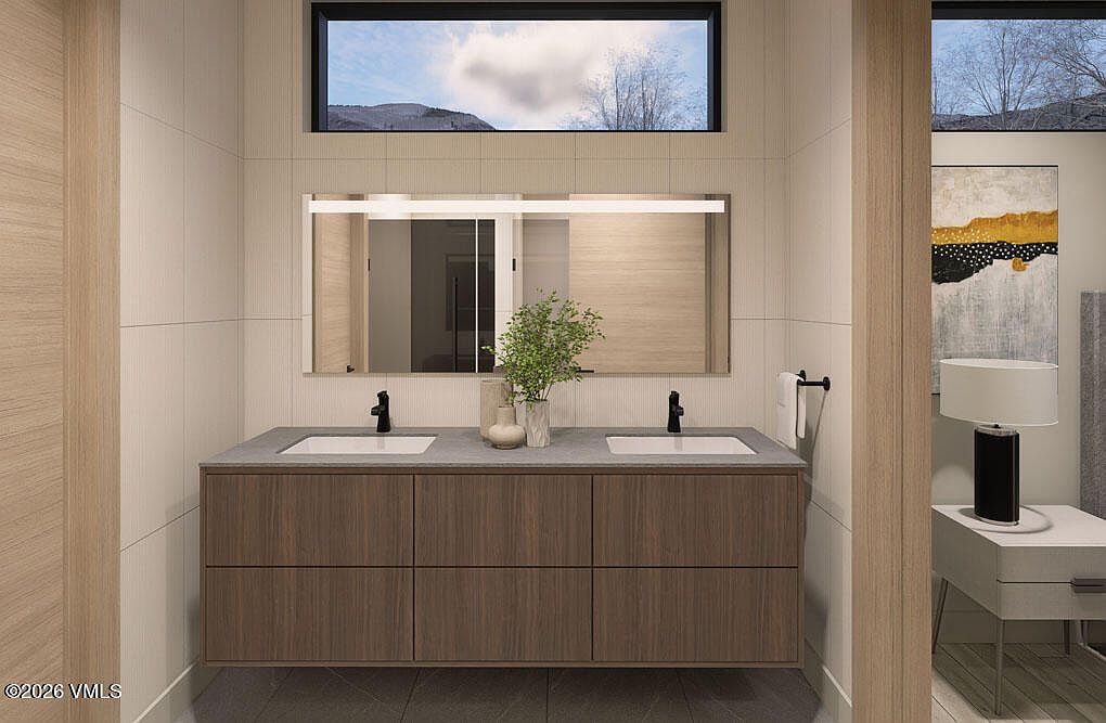 This is a well-lit primary bathroom featuring a double vanity with a dark wood finish and light gray countertop. A large mirror with integrated lighting hangs above the sinks, and a clerestory window provides natural light. The walls are clad in light-colored tiles, and the overall design is modern and minimalist.