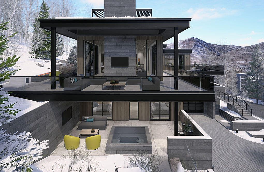 This image showcases a modern home's exterior, focusing on a multi-level patio/deck area. The upper level features comfortable outdoor seating with a glass railing offering views of the surrounding snow-covered landscape and mountains. Below, a lower patio includes a hot tub and additional seating, creating a luxurious outdoor living space.