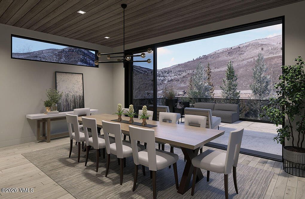 This is a dining room featuring a long wooden table with white chairs, set on a gray rug. A modern chandelier hangs above the table, and a large sliding glass door offers a view of a mountain landscape and an outdoor patio with seating. The room is well-lit and has a contemporary design.