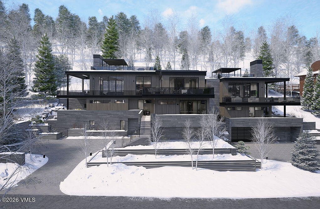 This is a front exterior view of a modern, multi-level home in a snowy, wooded setting. The house features a combination of stone and wood siding, with multiple balconies and large windows. The driveway and surrounding landscape are covered in snow, creating a serene and luxurious winter scene.