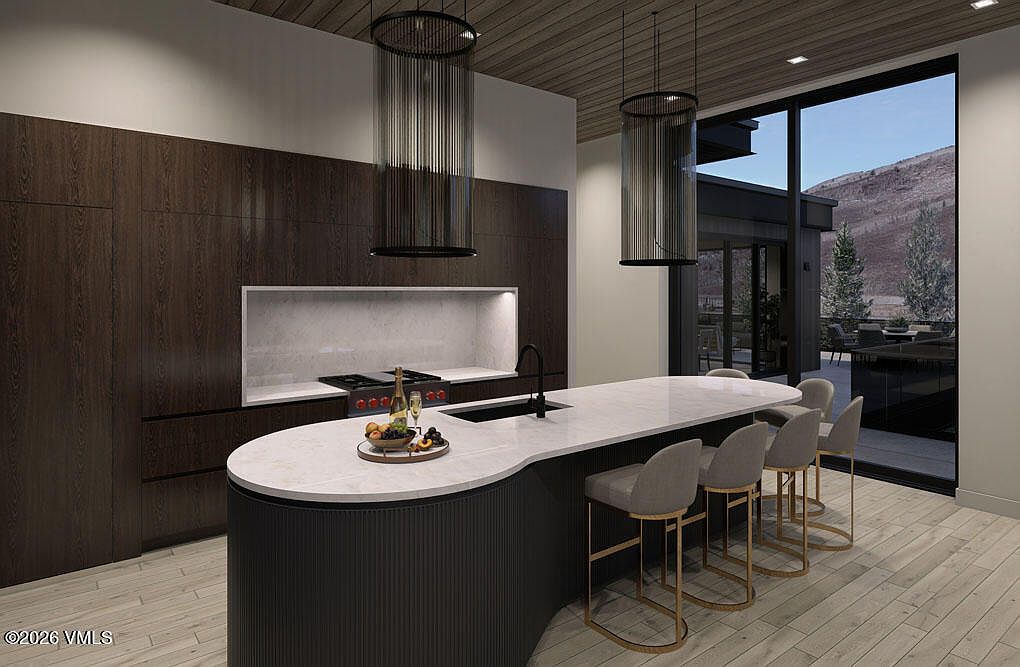 This is a modern kitchen featuring dark wood cabinetry and a large island with a white countertop and dark ribbed base. Two cylindrical pendant lights hang above the island, and a large window offers a view of the outdoor landscape. The kitchen has a sleek and contemporary design.