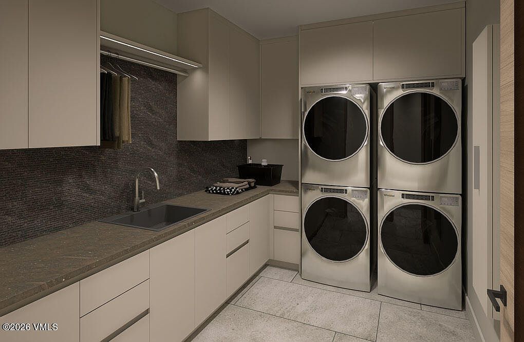 This is a well-organized laundry room featuring stacked washer and dryer units, light-colored cabinetry, and a dark countertop with a sink. The room has a modern and clean aesthetic, with a tiled backsplash and flooring. The perspective is from a medium distance, showcasing the layout and functionality of the space.