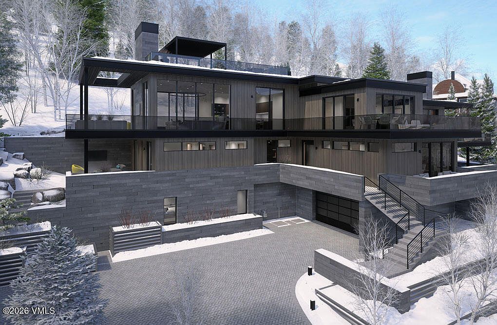 This image showcases the front exterior of a modern, multi-level home with a sophisticated design. The house features a combination of stone and wood siding, large windows, and multiple balconies, creating a luxurious and inviting appearance. The driveway is paved with interlocking stones, and the surrounding landscape is covered in snow, adding to the property's appeal.