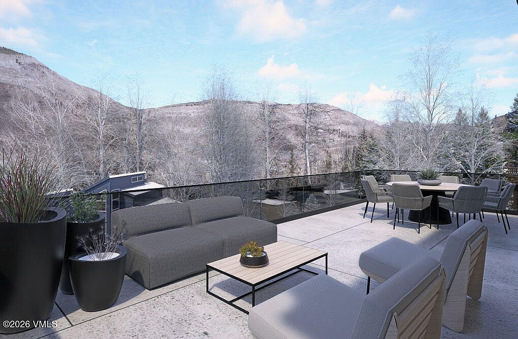 This image showcases a luxurious outdoor patio or deck area with modern furniture, including a gray sofa, chairs, and a dining table set, all arranged on a concrete surface. The space is surrounded by a glass railing, offering unobstructed views of a mountainous landscape with trees, some appearing snow-covered. The overall impression is one of sophisticated outdoor living with a focus on enjoying the natural scenery.