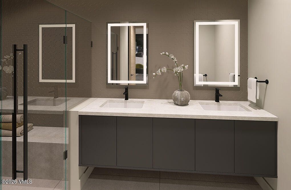 This is a modern primary bathroom featuring a double vanity with gray cabinetry and a white countertop. Two illuminated mirrors hang above the sinks, and a vase with flowers sits in the center of the countertop. A glass-enclosed shower is visible to the left, creating a clean and luxurious atmosphere.