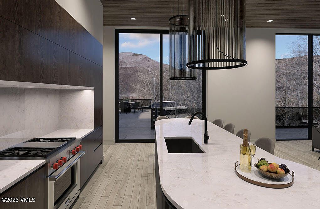 This is a modern kitchen with dark wood cabinetry and a large island featuring a white countertop. The kitchen is well-lit with pendant lighting and natural light from the sliding glass doors that lead to an outdoor patio with mountain views. The overall impression is luxurious and contemporary.