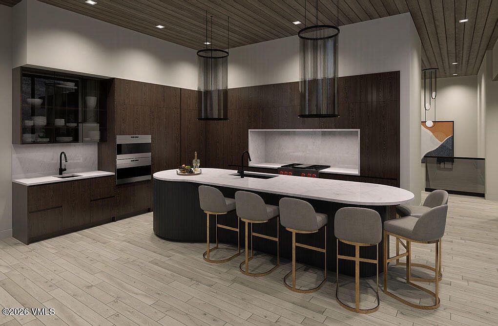 This is a modern kitchen with dark wood cabinetry, a large island with a white countertop, and four bar stools. The kitchen features stainless steel appliances and unique cylindrical pendant lighting. The overall impression is sleek and sophisticated, with a focus on clean lines and high-end finishes.