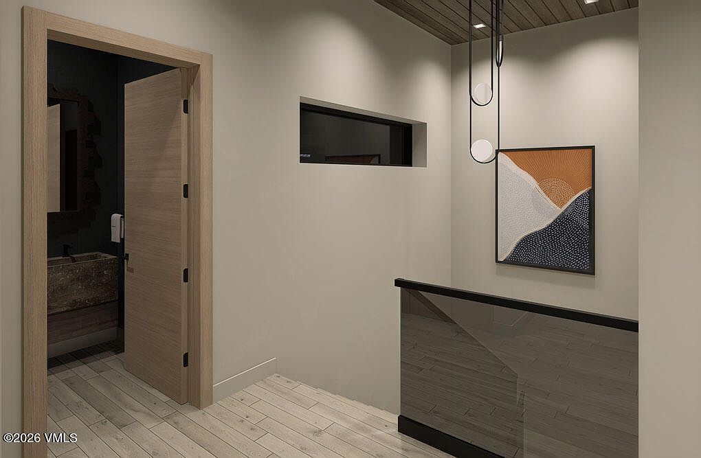 This interior shot showcases a modern hallway with light wood flooring and neutral-toned walls. A doorway leads to a bathroom, while a glass railing and an abstract art piece add contemporary touches. The space is well-lit, creating a clean and inviting atmosphere.