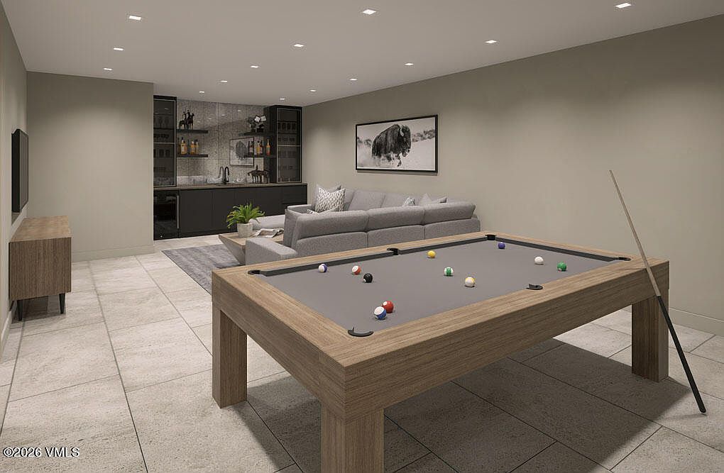This is a spacious basement featuring a pool table as the focal point, complemented by a comfortable seating area with a gray sectional sofa and a stylish bar area with dark cabinetry and open shelving. The room is well-lit with recessed lighting and features neutral-toned walls and tile flooring, creating a modern and inviting entertainment space.