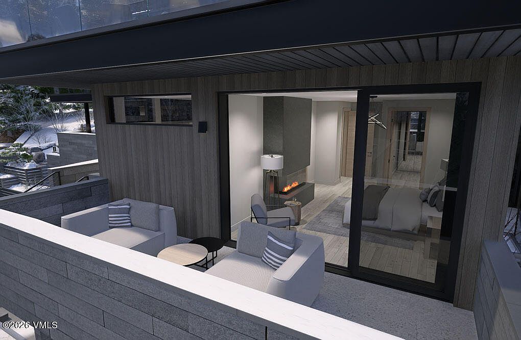 This image showcases a modern patio or balcony area with comfortable outdoor seating, including two light gray armchairs with striped pillows and a small round table. The space is connected to an interior room through large sliding glass doors, offering a glimpse of a contemporary bedroom with a fireplace. The overall design is sleek and inviting, blending indoor and outdoor living.